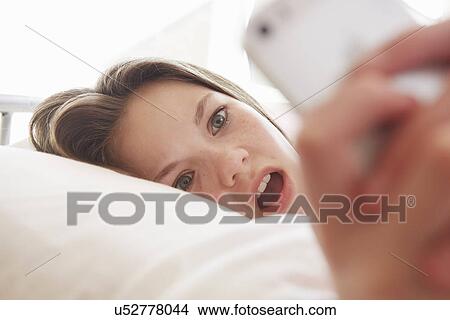 Surprised girl lying on bed reading smartphone message View Large Photo Image Picture - Surprised girl lying on bed reading smartphone message. Fotosearch