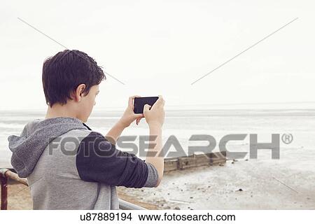 Picture - "Teenage boy photographing sea on smartphone, Southend on Sea, Essex, UK". Fotosearch