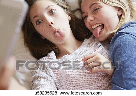 Two girls lying on bed sticking out tongues for smartphone selfie View Large Photo Image Stock Photo - Two girls lying on bed sticking out tongues for smartphone selfie. Fotosearch