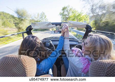 Stock Photo - "Two mature women, holding hands, in convertible car, rear view". Fotosearch