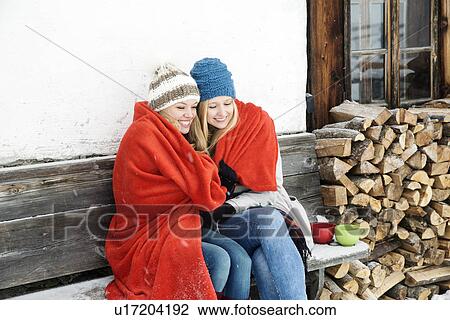 Two young female friends wrapped in red blanket sitting outside wooden cabin View Large Photo Image Stock Image - Two young female friends wrapped in red blanket sitting outside wooden cabin. Fotosearch