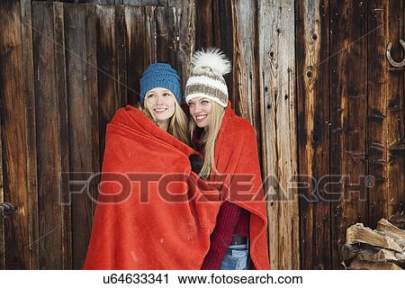 Stock Image - Two young female friends wrapped in red blanket outside wooden cabin. Fotosearch