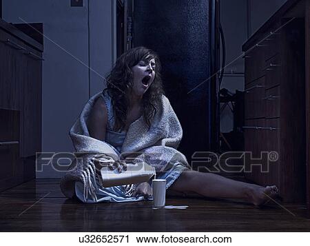 Stock Image - Yawning woman wrapped in blanket sitting on kitchen floor pouring milk at night. Fotosearch