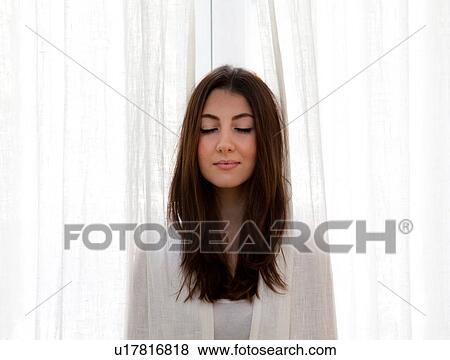 Stock Photo - "Young adult woman poking head out of curtains, eyes closed". Fotosearch