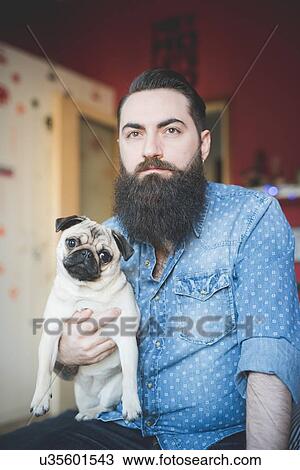 Stock Image - Young bearded man carrying dog in arms. Fotosearch
