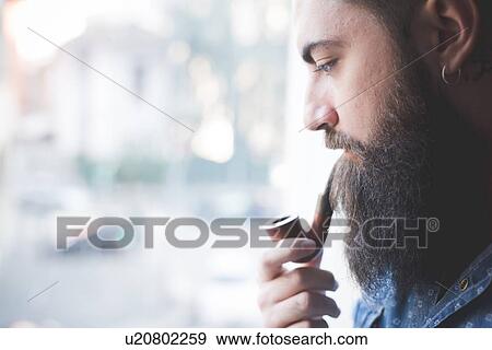 Stock Photo - Young bearded man smoking pipe by window. Fotosearch