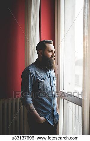 Young bearded man standing by window View Large Photo Image Stock Photograph - Young bearded man standing by window. Fotosearch