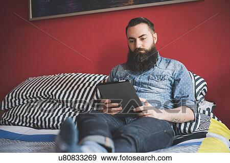 Young bearded man using digital tablet on bed View Large Photo Image Stock Photo - Young bearded man using digital tablet on bed. Fotosearch