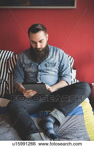 Stock Photograph - Young bearded man using digital tablet on bed. Fotosearch