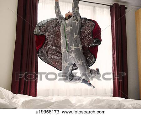 Stock Image - Young boy wearing cape jumping on bed. Fotosearch