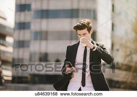 Young businessman drinking take away coffee whilst reading smartphone texts View Large Photo Image Stock Photo - Young businessman drinking take away coffee whilst reading smartphone texts. Fotosearch