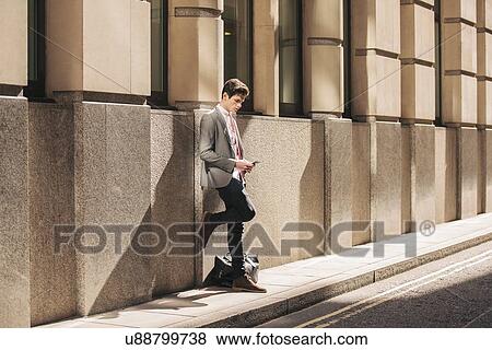 Young city businessman leaning against office building reading smartphone text View Large Photo Image Stock Photo - Young city businessman leaning against office building reading smartphone text. Fotosearch