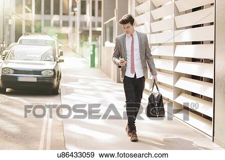Young city businessman reading smartphone texts whilst walking outside office View Large Photo Image Stock Photo - Young city businessman reading smartphone texts whilst walking outside office. Fotosearch