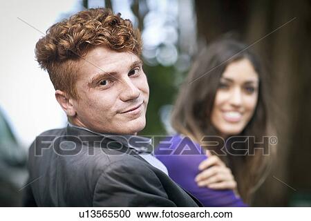 Stock Image - "Young couple sitting together, outdoors, looking at camera, smiling". Fotosearch