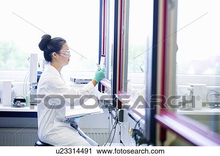 Stock Image - Young female scientist with notebook looking at sample in lab. Fotosearch