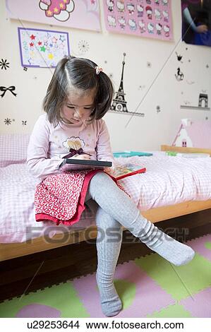 Picture - Young girl sitting on bed looking at DVD cover. Fotosearch