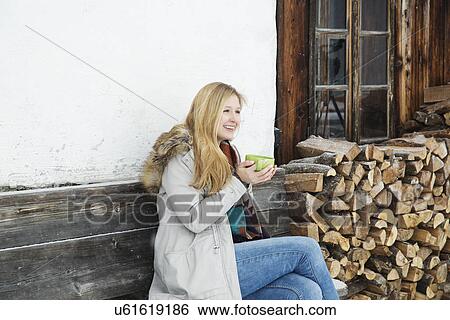 Stock Photograph - Young woman sitting outside wood cabin drinking coffee. Fotosearch