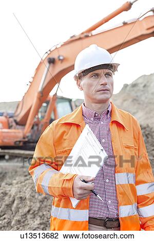 Stock Image - Architect looking away while holding clipboard at construction site. Fotosearch