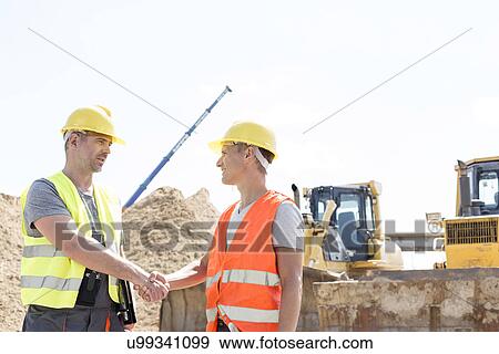 Stock Photo - Architects shaking hands at construction site against clear sky. Fotosearch