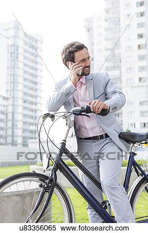 Stock Photography - Businessman answering mobile phone while standing with bicycle outdoors. Fotosearch