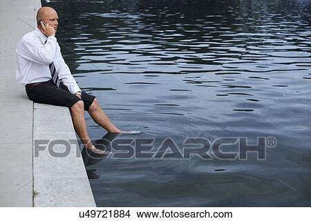 Picture - Businessman sitting on wall by lake, using mobile phone. Fotosearch