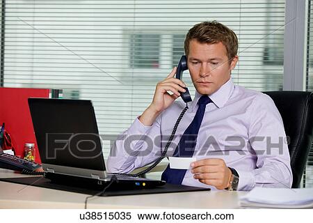 Stock Photography - Businessman talking on telephone and looking at card in office. Fotosearch