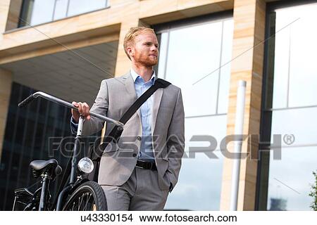 Picture - Businessman with bicycle looking away while standing outside office building. Fotosearch