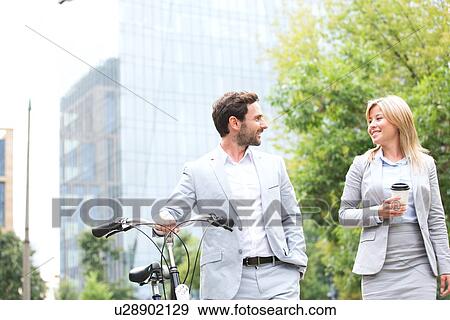 Businesspeople with bicycle and disposable cup conversing while walking outdoors View Large Photo Image Stock Photo - Businesspeople with bicycle and disposable cup conversing while walking outdoors. Fotosearch