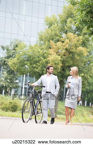 Stock Image - Businesspeople with bicycle conversing while walking on street. Fotosearch