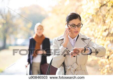 Businesswoman checking time while colleague standing in background at park View Large Photo Image Stock Image - Businesswoman checking time while colleague standing in background at park. Fotosearch