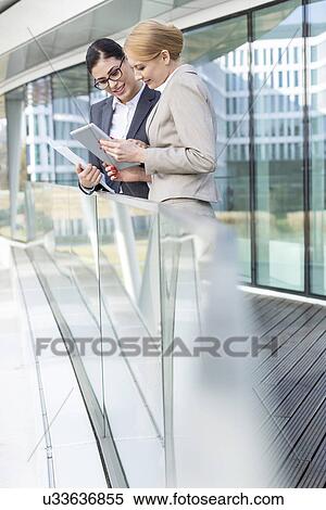 Stock Photography - Businesswomen using digital tablet by glass railing. Fotosearch