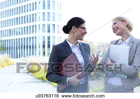 Cheerful businesswomen conversing while holding folder and laptop outside office building View Large Photo Image Stock Photo - Cheerful businesswomen conversing while holding folder and laptop outside office building. Fotosearch