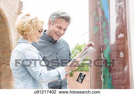 Stock Image - Cheerful middle-aged couple reading map in city. Fotosearch