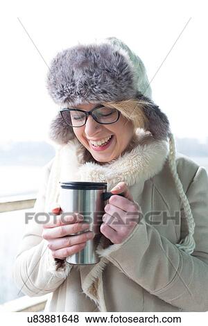 Stock Photo - Cheerful woman in warm clothing holding insulated drink container. Fotosearch
