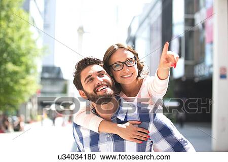 Cheerful woman pointing away while enjoying piggyback ride on man in city View Large Photo Image Stock Image - Cheerful woman pointing away while enjoying piggyback ride on man in city. Fotosearch