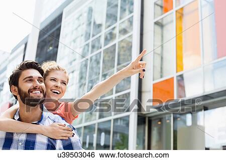 Stock Photograph - Cheerful woman showing something to man while enjoying piggyback ride in city. Fotosearch