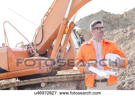 Stock Photo - Confident architect holding hardhat and blueprints at construction site. Fotosearch