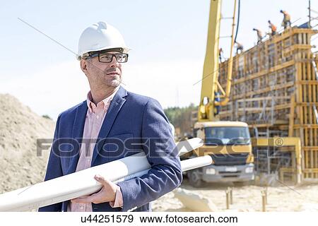 Stock Photo - Confident architect holding rolled up blueprints at construction site. Fotosearch