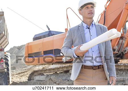 Stock Image - Confident architect looking away while holding blueprints at construction site. Fotosearch