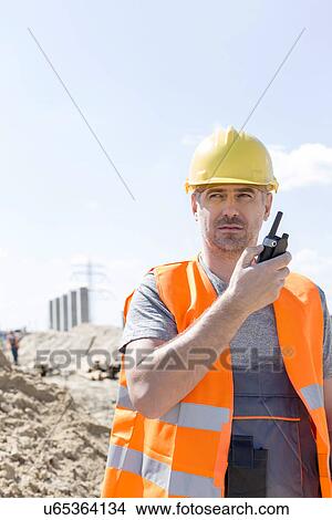 Confident supervisor using walkie-talkie at construction site against sky View Large Photo Image Picture - Confident supervisor using walkie-talkie at construction site against sky. Fotosearch