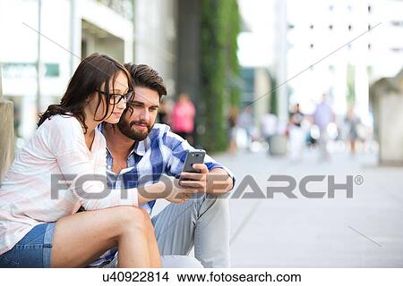 Picture - Couple using smart phone together while sitting on steps in city. Fotosearch