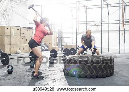 Stock Photo - Dedicated man and woman hitting tire with sledgehammer in crossfit gym. Fotosearch