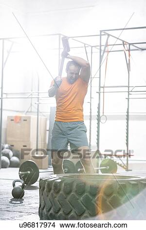 Picture - Determined man hitting tire with sledgehammer in crossfit gym. Fotosearch