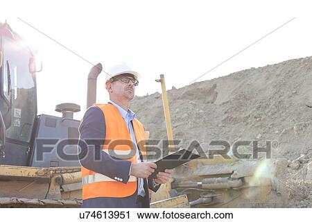 Stock Image - Engineer looking away while holding clipboard at construction site. Fotosearch