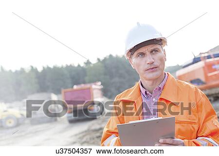 Stock Photo - Engineer looking away while holding clipboard against clear sky. Fotosearch