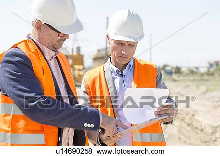 Stock Photo - Engineers examining documents on clipboard at construction site against clear sky. Fotosearch