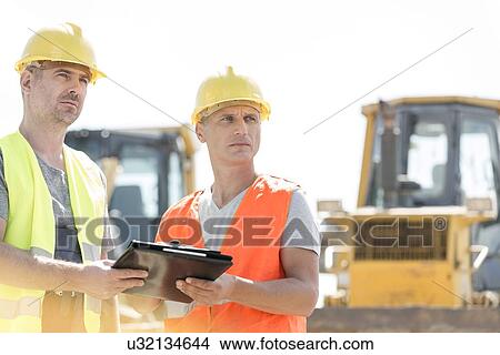 Picture - Engineers looking away while holding clipboard at construction site against clear sky. Fotosearch