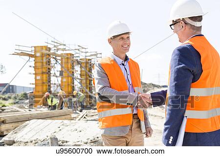 Stock Image - Engineers shaking hands at construction site against clear sky. Fotosearch