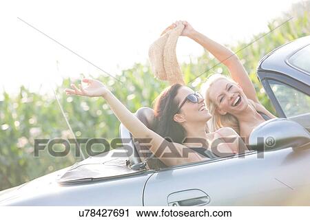 Stock Image - Excited female friends enjoying road trip in convertible on sunny day. Fotosearch