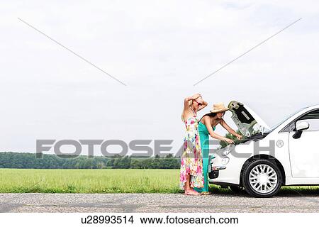 Picture - Female friends examining broken down car on country road against clear sky. Fotosearch
